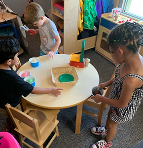 Three students playing with Play Doh around a table in a classroom